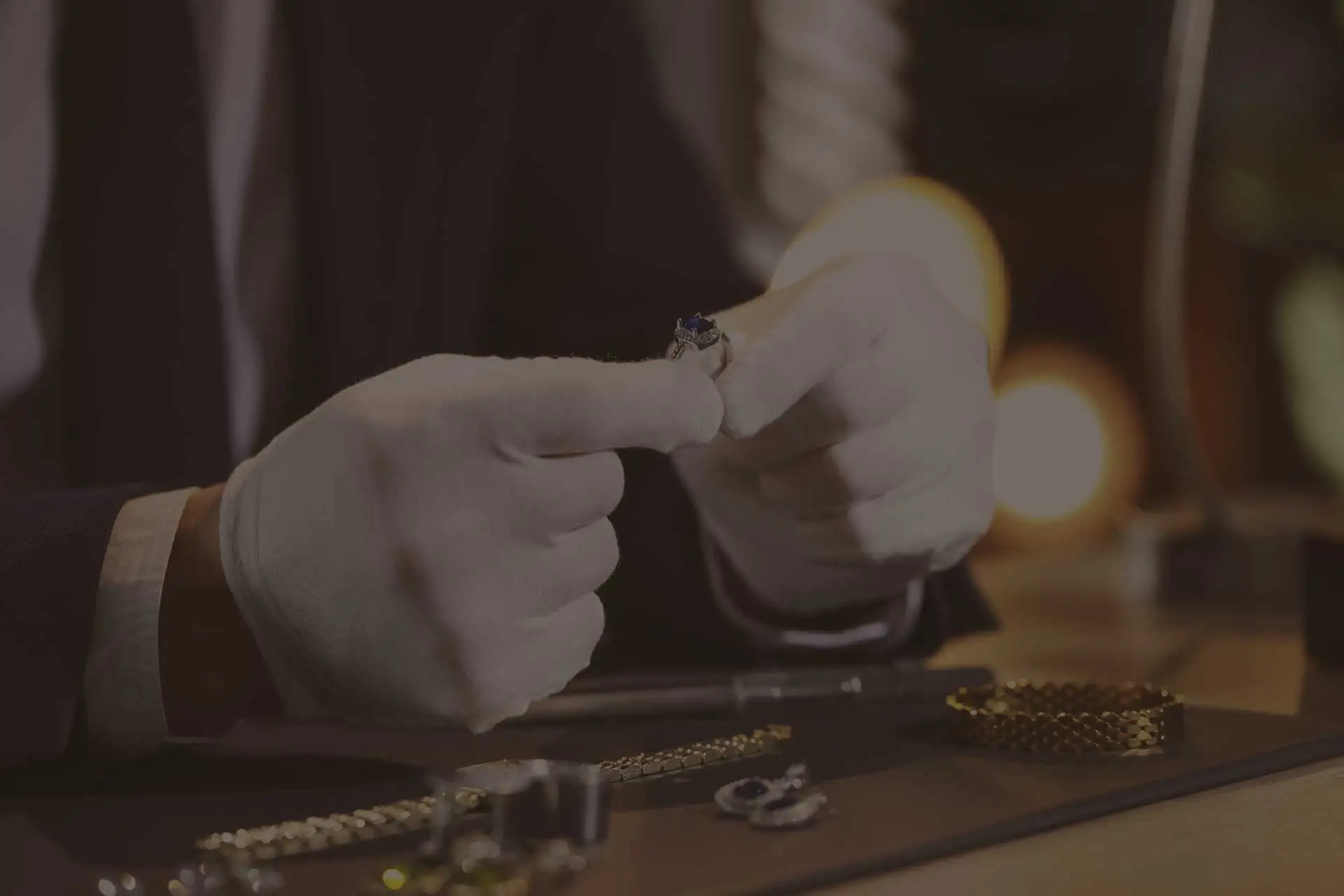 Experte in Anzug und weißen Handschuhen begutachtet einen Ring mit blauem Edelstein über einem Tisch mit Goldschmuck.
