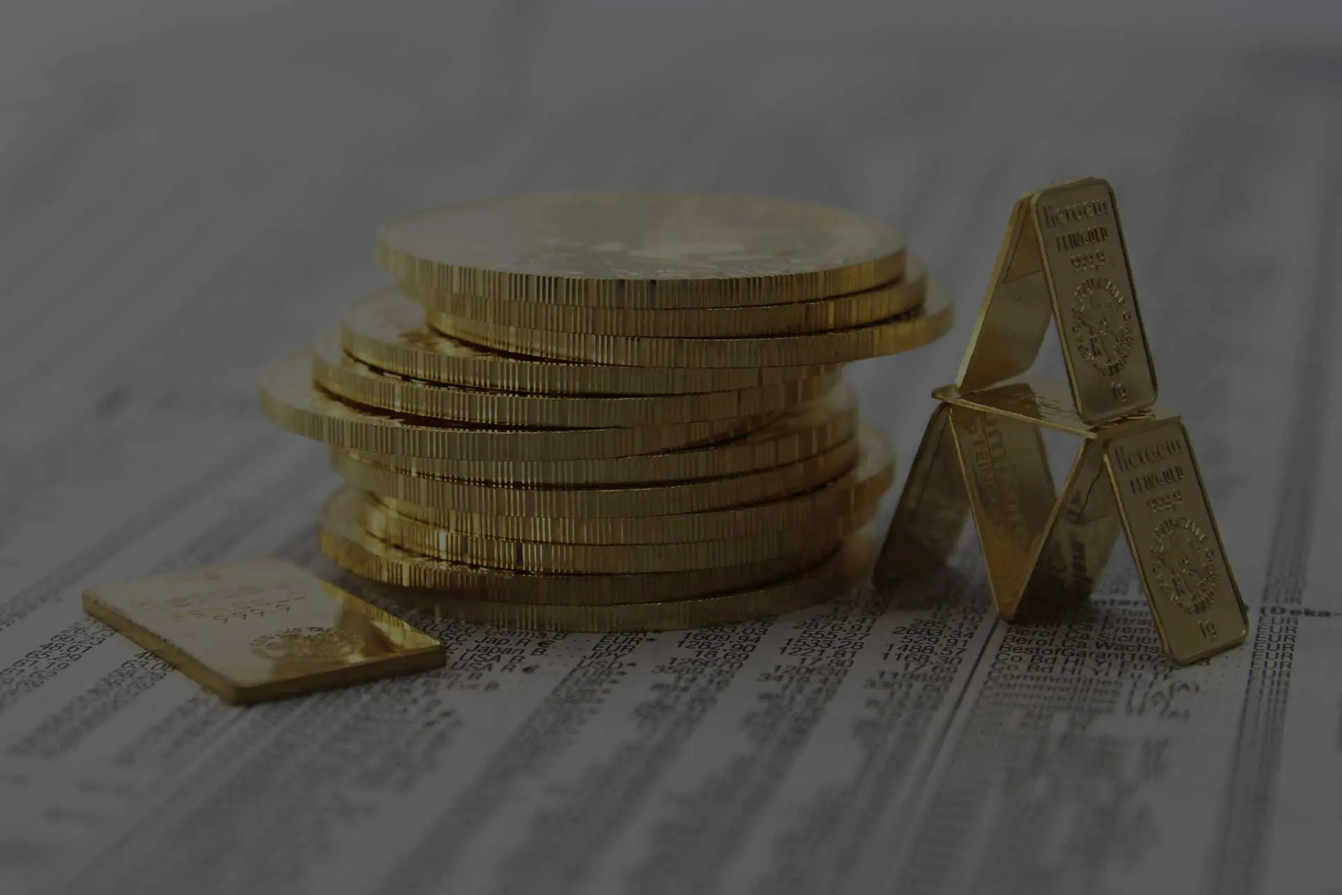 Stapel aus Goldmünzen und kleine Goldbarren von Umicore und Heraeus als Kartenhaus auf Kursnotierungen einer Zeitung.